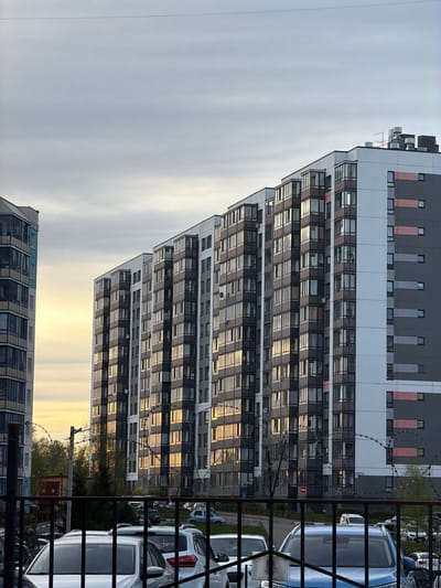 The image depicts a multi-story apartment building in Pavlovo, Russia. The building has a modern design with alternating grey and white sections and numerous windows that reflect the sunlight. In the foreground, there is a metal fence with barbed wire. Parked cars are visible behind the fence. The sky is overcast.