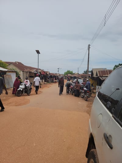 The image depicts a street scene in Tangu, Nigeria, likely a marketplace or a busy thoroughfare. The ground is covered with reddish-brown earth, and a paved section of the road runs through the center. Along the sides of the road are numerous small shops and structures with corrugated metal roofs, indicating a commercial area.

Several people are visible, engaged in various activities. Some are walking, while others are gathered around motorcycles, suggesting transportation or delivery services are active. There are at least five motorcycles visible in the image, some with riders. One motorcycle in the foreground is white with black accents.

A white van is partially visible on the right side of the frame, with its side and rear quarter panel in view. Its presence suggests that motor vehicles are common in this location.

The sky is overcast, with a light gray appearance, suggesting it might be a cloudy day. There are power lines running across the scene, supported by wooden poles, and a solar-powered street lamp is also visible.

The overall impression is of a vibrant, active street in a Nigerian town or city, characterized by a mix of commercial activity and local life.