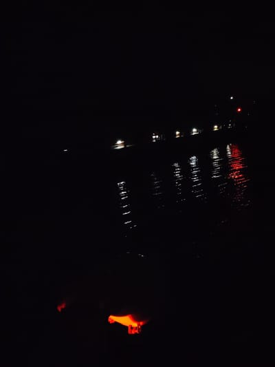 The image appears to be taken at night in Pruzhinino, Russia, near a body of water. There are lights visible across the water, including white and red lights which could indicate buildings or traffic. Reflections of these lights are visible on the water surface. In the foreground, there is a blurred orange glow, potentially from an object or source of light that is close to the camera. Due to the darkness and limited visibility, further details are difficult to determine with certainty.