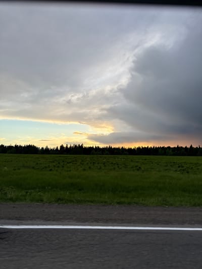 The image shows a landscape with a road in the foreground, followed by a green field, a line of trees, and a dramatic sky with dark storm clouds and a glimpse of golden light. The road has a white line marking its edge. There are no people visible in the image. The location is Meshcheryaki, Russia.