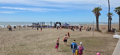 This image depicts a beach scene in Benicàssim, Spain. The sky is blue with scattered clouds, and the sea is visible in the background with several ships. A sandy beach stretches across the foreground and midground.
There is a gathering of people near a black inflatable arch labeled "Arena," presumably an event setup for a swimming or triathlon event. Some people are wearing pink swim caps and carrying safety floats, indicating they are participants. There are tents, flags, and umbrellas scattered around the event area.
In the foreground, several individuals are walking away from the event area. Some are shirtless men carrying safety floats; others wear backpacks. Palm trees line the right side of the image, and a building is partially visible in the bottom right corner. Overall, the scene appears to be a sunny day at the beach with an event taking place.