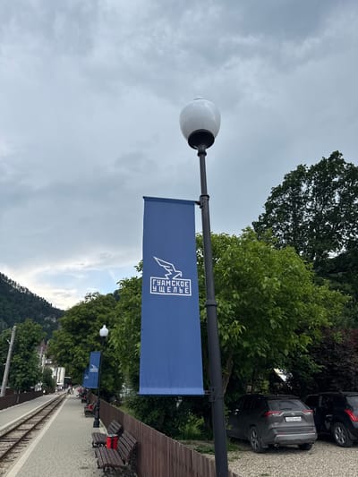 A blue banner with white text and a bird silhouette hangs from a lamppost. The text on the banner appears to read "ГУАМСКОЕ ЩЕЛЬЕ", which translates to "Guam Gorge". In the background, there are trees, a gravel parking lot, and parked cars. One car has a license plate that reads "C070XY 34". A wooden walkway with benches and a set of train tracks are visible in the distance. The scene appears to be a public area, possibly a park or tourist attraction, in Guamka, Russia. The sky is overcast, suggesting it might be a cloudy or late afternoon.