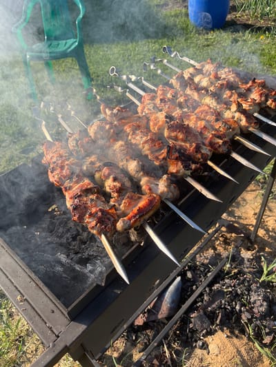 The image shows a barbecue grill with multiple metal skewers laden with grilling meat, likely shashlik. The skewers are arranged across the top of the grill, which appears to be charcoal-fueled, judging by the smoldering coals underneath and the rising smoke. The surrounding environment consists of grass and some sandy, bare earth. A blue container and a green chair are visible in the periphery. The location is identified as Сапуголи, Russia.