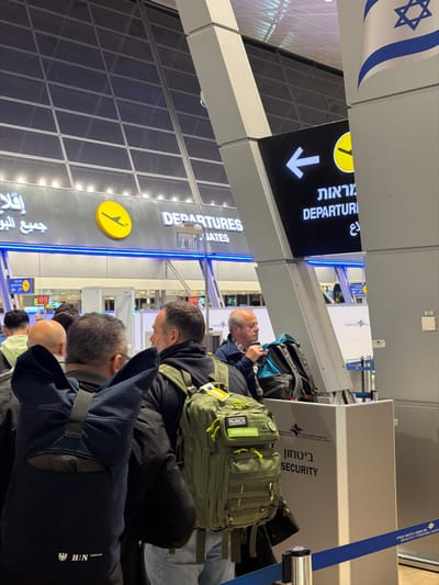 At an airport, travelers line up near a "Departures" sign, written in English, Arabic, and Hebrew. There is also a sign for security. A man with a backpack with a "Munich Sports" patch on it is close to the head of the line. A person with a bag with the logo "HIN" is close to the front. The Israeli flag is on the ceiling.