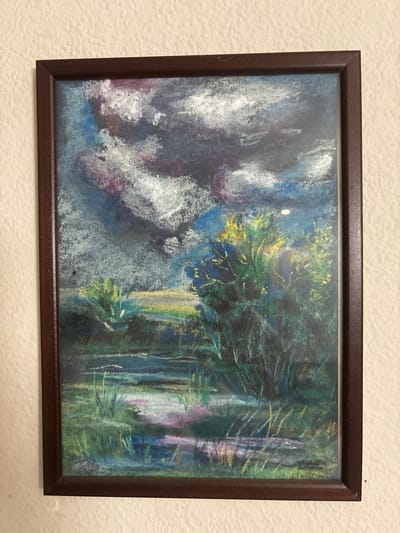 The image shows a framed pastel drawing of a landscape. The drawing depicts a dark, cloudy sky with hints of blue and purple, above a scene of marshy land with water and vegetation. Tall reeds and grasses are visible along the water's edge, and a cluster of trees with yellow and green foliage is prominent on the right side of the drawing. The water reflects the colors of the sky and surrounding landscape. The drawing is framed in a dark brown wooden frame and hangs on a textured, off-white wall. The location of Svkh Maslovskiy 1 Otdeleniye, Russia, is not depicted in the image.