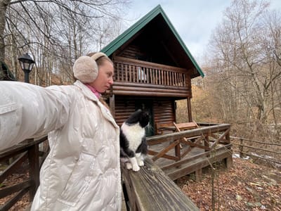 The image captures an outdoor scene in Crkvine, Montenegro, featuring an adult female and a domestic cat on a wooden deck in front of a log cabin. The female, positioned on the left side of the frame, is seen in profile, facing left, with her left arm extended, holding an unseen device consistent with taking a self-portrait. She wears a light-colored, likely beige or cream, shiny puffer jacket with a fleece-lined collar, a pink garment visible underneath, and large, fluffy beige earmuffs. Her light-colored hair is partially visible.
To her right, a black and white domestic cat sits upright on a sturdy wooden railing, facing right, with its head turned away from the camera. The cat exhibits a white chest, paws, and facial markings against a primarily black body.
In the background, a two-story wooden log cabin with a dark green A-frame roof is prominently featured. The cabin has a second-floor balcony with a wooden railing and a first-floor deck where a wooden bench is visible. The structure is constructed from dark brown logs. Surrounding the cabin and subjects is a dense forest of deciduous trees with bare branches, suggesting late autumn or winter. The ground is covered with fallen brown and orange leaves. An old-fashioned black outdoor lamp post is visible in the upper left portion of the image, behind the female. The sky is uniformly overcast and light grey.
