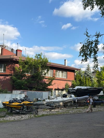 The image depicts a waterfront scene in Povedniki, Russia, on a sunny day. The foreground shows a paved path next to a grassy area where several watercraft are parked on trailers. Two yellow jet skis are prominently displayed, each with luggage strapped to the seats. Behind them, a dark-hulled boat with "KOCATKA" and "TA 3698RUS50" written on its side is visible, along with other boats and trailers. A person in beige pants and a white top is seen leaning against the dark boat.
In the background, a red brick building with white-framed windows stands behind a lush green tree. The building appears to be a house or a small commercial establishment. The sky is a bright blue with scattered white clouds, suggesting fair weather. The overall setting suggests a recreational area, likely near a lake or river, where water sports equipment is stored and accessed. The presence of the jet skis and boats indicates a focus on water-related activities. The image has a casual, summery feel.