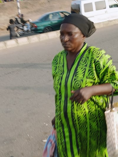 The scene is located in the city, near Kungulu School, Zambia. An adult female is positioned in the foreground, walking from right to left across a paved road surface. She wears a black headscarf and a loose-fitting, vibrant green garment adorned with black vertical stripes and circular patterns. Her expression is neutral. She carries a folded, multi-colored fabric object in her left hand and a light-colored, patterned bag in her right hand. In the blurred midground, a low concrete barrier separates the foreground pavement from a street featuring vehicular traffic. A dark green sedan is visible, along with a partially obscured white van further back. To the left of the sedan, a motorcycle carries two individuals. Another person in dark clothing is partially visible walking on the far left. A light-colored pole is also present on the left side of the frame. The environment indicates an active urban street scene during daylight hours.