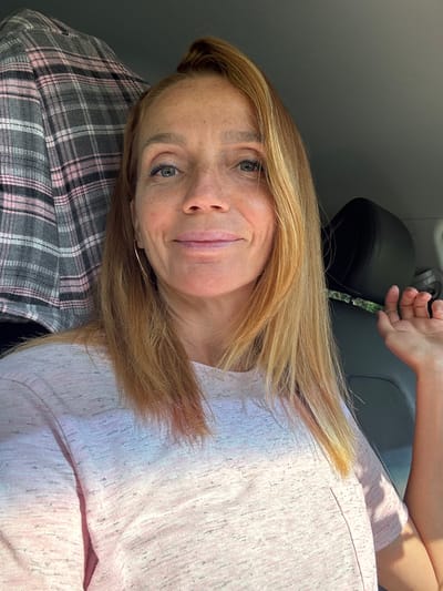 A blonde woman with fair skin and freckles is photographed from the chest up, sitting in what appears to be a vehicle. She is looking directly at the camera with a slight smile. Her hair is long and blonde, pulled back on top of her head, with some strands falling around her face. She is wearing a light pink t-shirt with a textured pattern. To her left, a gray and pink plaid blanket or piece of fabric is visible. To her right, a black car headrest is seen, and her right hand is raised, palm facing away from the camera, with her fingers slightly spread. The lighting suggests it is daytime, and the overall scene is casual and personal. The image does not contain any elements that would allow for specific identification of the location within Portovoe, Russia.