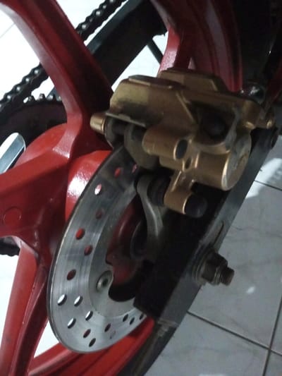 A close-up shows the rear wheel and braking system of a motorcycle. A red, five-spoked wheel is visible with a black chain and sprocket. A silver disc brake is mounted to the wheel's hub. A gold-colored brake caliper is attached to the motorcycle's black swingarm. The swingarm is secured with a nut and bolt. The background shows a white tiled floor and part of a wall.