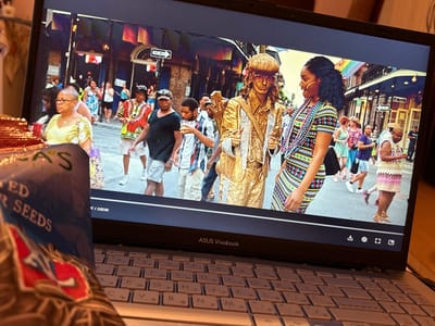 An ASUS Vivobook laptop with a Cyrillic keyboard is open on an indoor surface in Kastel, Bulgaria. In the immediate foreground, a brown and blue packet, partially visible, indicates "SUNFLOWER SEEDS." The laptop screen displays a video of a busy street scene. In the video's foreground, a human statue covered in gold paint, wearing a suit, hat, and sunglasses, stands on the left. To the right of the statue, a Black woman with braided hair, adorned with multiple beaded necklaces, wears a colorful striped dress and looks towards the statue. The background of the video shows a narrow city street lined with multi-story buildings, featuring a "ONE WAY" sign and a rainbow flag. Numerous pedestrians of varied demographics are walking along the street.