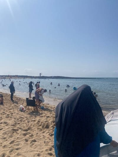 The image depicts a busy beach scene in Harhoura, Morocco, on a clear, sunny day. The foreground is dominated by sandy beach with footprints. In the middle ground, a diverse group of people are enjoying the water and beach activities. Several individuals are wading or swimming in the calm sea. Some are seen interacting with each other, while others are engaged in solo activities. Beach chairs and bags are scattered on the sand, indicating a leisurely day. In the background, a long stone jetty stretches into the sea, and a lifeguard tower is visible atop it, suggesting an organized and safe beach environment. The sky is a clear, pale blue, indicating good weather conditions. The overall impression is one of a lively and popular coastal resort.