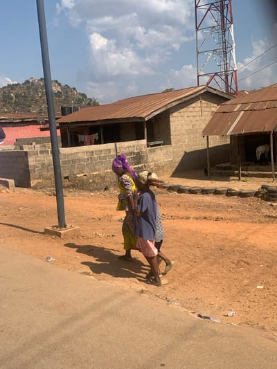 The image depicts a daytime scene in Egbedi, Nigeria, under a bright blue sky with scattered white cumulus clouds. Three individuals are walking from right to left on a reddish-brown dirt path that borders a darker, likely paved, surface in the bottom left foreground. The lead figure, an adult female, wears a yellow-green patterned top and a purple headwrap. Behind her, two children follow; one wears a dark blue t-shirt and light pink shorts, the other a striped top. All three cast distinct shadows to their left.
In the mid-ground, a dark grey utility pole with a concrete base stands on the left. Behind the path, multiple single-story buildings constructed with unpainted concrete blocks and topped with rusty corrugated metal roofs are present. Clothes are visible hanging outside the left-most building, which also has a black water tank on its roof. To the far right, a red and white lattice-patterned communications tower rises into the sky. A smaller, open-front structure with a corrugated metal roof, appearing to be an animal pen, houses a white goat-like animal near its steps. Discarded tires are arranged along the edge of the dirt path near this pen, and a small, dark animal is near the pen's entrance. The background features rocky hills under the clear sky.