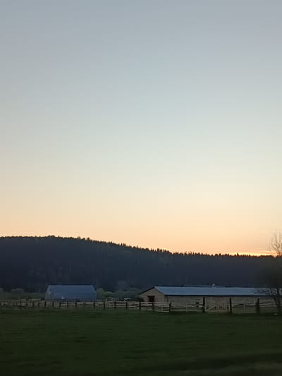 The image shows a rural landscape in Ust-Kishert, Russia, at sunset. A dark, densely forested hill forms the horizon. In the foreground, a grassy field stretches towards a wooden fence. Two barns are visible behind the fence: one with a gray roof and walls and the other barn is stone or masonry construction with a silver roof. The sky transitions from pale blue to orange near the horizon.