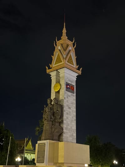 This is a nighttime, full-frame, low-angle shot of the Independence Monument in Khan Daun Penh, Cambodia. The monument, a tall, white, stone structure with a tiered, gold-trimmed roof and spire, is centrally located against a dark sky. Two flags are affixed to the monument's facade: one with blue and red stripes and a depiction of Angkor Wat, and another, smaller red flag with a gold emblem. At the base of the monument, in front of the stone tower, are three bronze statues of individuals standing closely together. The statues appear to be a man and two women, with the man on the right holding a rifle. Below the statues is a plaque with text. In the lower left of the frame, the top of a temple with a golden roof is visible, as are trees and lampposts. The scene is illuminated by artificial lights, casting a warm glow on the monument and surrounding structures.