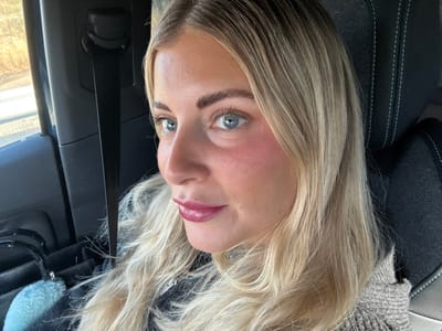 A blonde female, with blue eyes and reddened cheeks, is depicted from the mid-chest up, seated in the passenger side of a vehicle in Meyreuil, France. She wears dark red lipstick and has long, wavy blonde hair. Her attire consists of a dark zippered garment under a light brown or beige knitted outer layer. A black car seat with white stitching is visible behind her head and to her right. A black seatbelt is positioned vertically to her left. Through the car window to her left, blurred brown and green foliage suggests an outdoor environment. A light blue fluffy keychain is visible near the bottom left of the frame, attached to an unseen object. The subject is looking slightly to her left, without direct interaction with any other visible entities.
