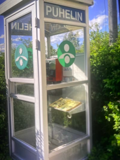 A public phone booth, labeled "PUHELIN" at the top, stands in a green, outdoor environment. The booth is constructed of white metal and glass, with a single glass door. Inside, a red and black telephone machine is visible. Below the telephone, a yellow and green item, possibly a book or pamphlet, rests on a shelf. Two green circular logos, each depicting a stylized human figure, are affixed to the glass on the left side of the booth. The background consists of lush green trees and a bright blue sky with white clouds. The scene appears to be set in a semi-rural or suburban area, potentially a park or a quiet street. The image's resolution and focus suggest it may have been taken with a mobile phone camera. The overall impression is one of an aging but functional piece of public infrastructure in a natural setting.