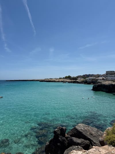 This image presents a coastal scene in Monopoli, Italy, under a clear, bright blue sky containing several faint, wispy contrails, two of which are prominent in the upper left quadrant. The sea exhibits distinct coloration, appearing turquoise and transparent in the foreground and shallower areas, revealing a rocky seabed, and transitioning to a darker blue further towards the horizon. The water surface shows numerous small ripples reflecting sunlight.

The left side of the frame features a dark, rocky promontory extending into the sea, topped with sparse, light-colored vegetation. A small, dark-colored inflatable boat or raft is partially visible in the water near the far left edge.

The right side of the image is characterized by a rocky shoreline. In the immediate foreground, large, dark grey and black rock formations are visible, with a patch of green vegetation on the extreme lower right. Further along the coastline to the right, a town or settlement is built directly on and above the cliffs. This includes various multi-story buildings in shades of white, beige, blue, and grey. A significant white building with an outdoor terrace and umbrellas, possibly a restaurant or cafe, is situated prominently at the water's edge.

Two individuals are visible in the turquoise water on the right side, appearing to be swimming or floating. One person is closer to the foreground, with their head and shoulders above the water, facing away from the viewer. The second person is slightly further back, also with their head and shoulders visible, and appears to be utilizing a small white flotation device. No discernible interaction between the individuals is evident.