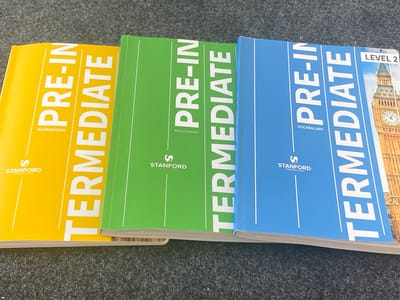 Three books titled "PRE-INTERMEDIATE" are lying on a dark gray, textured surface that appears to be carpet. The books are arranged diagonally from left to right, with the leftmost book being yellow, the middle book green, and the rightmost book blue. The yellow book has "WORKBOOK" printed below the title. The green book has "READING BOOK" printed below the title. The blue book has "VOCABULARY" printed below the title, and "LEVEL 2" is printed in the top right corner. Each book features the "Stanford" logo. The background shows a portion of a building with a clock tower, resembling Big Ben in London, on the right side of the image. There are no people present in the image.