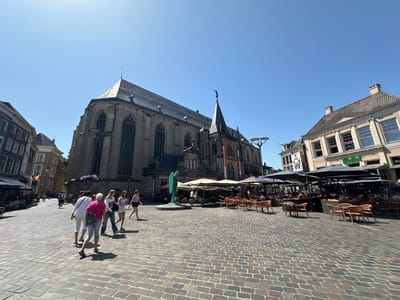 This media file captures a sunlit day in a European city square, likely Zwolle, Netherlands. The scene is dominated by a grand, Gothic-style church with a tall steeple, its stone facade etched with the inscription "ANNO 1520." In front of the church, a modern abstract sculpture of a stylized green human figure stands prominently on a circular base.

The square is paved with cobblestones, and scattered throughout are tables and chairs belonging to outdoor cafes and restaurants, many of them covered by large umbrellas. Several groups of people are visible: a cluster of four women walking together in the foreground, dressed in casual summer attire, with one in a pink top and jeans carrying a dark grey bag. In the background, patrons are seated at the outdoor dining areas, enjoying the weather. A person is seen riding a bicycle on the periphery of the square.

The time of day appears to be midday, given the bright sunlight and strong shadows cast by the people and objects. The sky is a clear, vibrant blue, indicating pleasant weather. The architecture of the surrounding buildings, with their multi-paned windows and tiled roofs, further suggests a European setting. One building displays a sign that reads "DE HARMONIE," with "Grolsch" visible on another nearby establishment. The overall atmosphere is one of a lively, communal gathering space on a beautiful summer day.