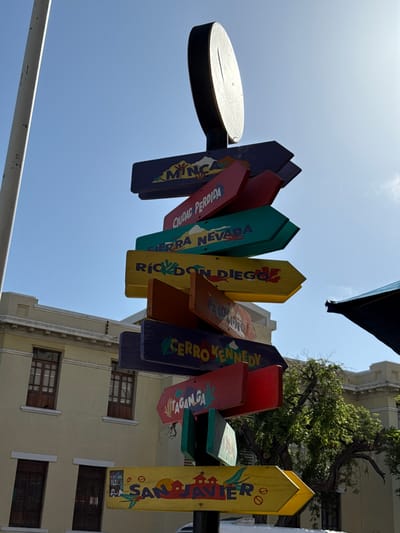This media file captures a collection of colorful, hand-painted wooden signs pointing in various directions, mounted on a pole. The signs are angled and overlap each other, creating a dynamic visual. Each sign appears to list a different destination, with some names partially visible.

The subjects are primarily the signs themselves, which are depicted in detail, showcasing their varied colors and the hand-painted text. The scene is an outdoor setting, with a bright blue sky dominating the upper portion of the image, suggesting a clear day. The lighting indicates it is daytime, likely mid-morning or afternoon due to the bright sun casting some reflections on the top sign.

Notable details include the design of the signs, which are shaped like arrows and have a rustic, handcrafted appearance. Some of the visible text on the signs includes "CIUDAD PERDIDA," "NEVADA," "RIO DI D," "GUAYAMA," "CERRO KENED," and "SAN JAVIER." The presence of these names suggests a tourist destination or a location with multiple points of interest. In the background, the upper floors of a light-colored building with visible windows can be seen, hinting at an urban or semi-urban environment. The overall impression is one of a vibrant and perhaps slightly whimsical tourist landmark. The context provided states the location is Santa Marta, Colombia, which aligns with the likely tropical or subtropical destination implied by the signs.