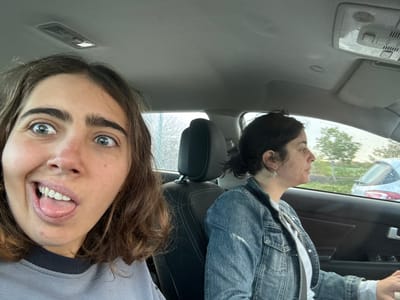 Two females are depicted inside a vehicle in the city of Nes Ziona, Israel. The individual in the front passenger seat, a young female with brown hair and light eyes, is positioned in the foreground, facing the camera with a playful expression: eyes widened and tongue slightly extended. She wears a grey sweatshirt. In the driver's seat, an adult female with dark hair pulled back, wearing a denim jacket over a white top, is visible in profile, looking forward. Her right hand rests on what appears to be the steering wheel or armrest. Raindrops are visible on the side window, and the exterior environment is blurred, showing green foliage and a partial view of another light-colored vehicle in the background. The car's interior features a grey headliner and dark seating.