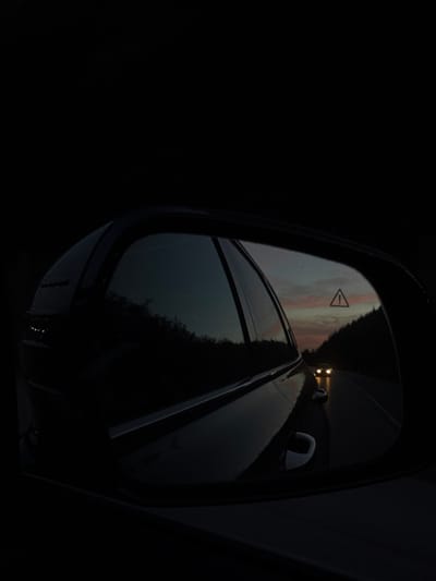 The image depicts a forensic view from a vehicle's passenger-side wing mirror at dusk in Gorna Vasilitsa, Bulgaria. The mirror's dark, glossy casing frames a reflection of a two-lane road stretching into the distance under a twilight sky featuring faint orange and pink hues. Dense, dark foliage or hills line both sides of the road. Two distinct, illuminated headlights from an approaching vehicle are visible in the center of the reflection. Above the approaching vehicle, an illuminated triangular warning symbol containing an exclamation mark is present in the upper-center of the mirror's reflection. The immediate environment outside the mirror's reflection is obscured by deep darkness.