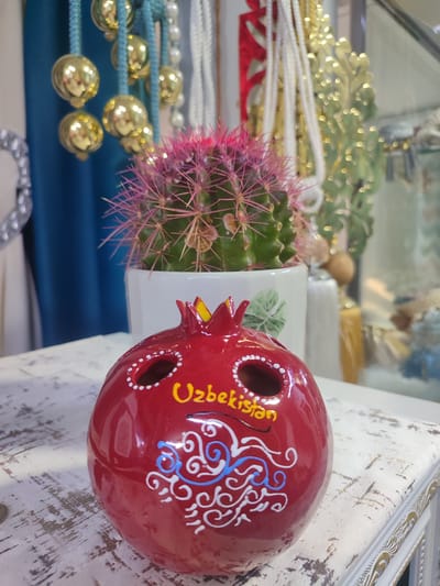 In Batken City, Kyrgyzstan, a close-up shot captures a red, pomegranate-shaped ceramic ornament with decorative white and blue swirls. Yellow lettering on the ornament reads "Uzbekistan." A green cactus with pink spines in a white pot is positioned behind the ornament. The background features blurred decorative elements, including gold beads, pearl strings, and what appear to be fabric drapes in shades of blue and white. The items are placed on a distressed white surface with visible wood grain.