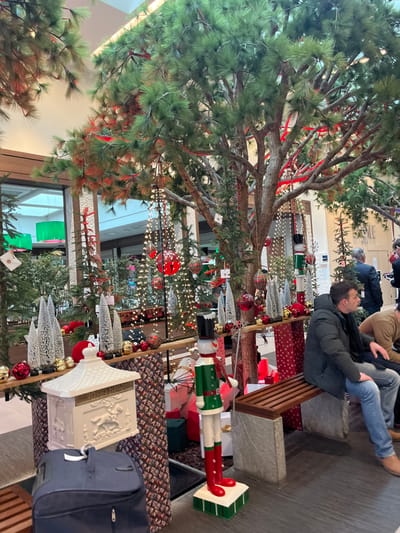 This image depicts an indoor commercial space in Ribeauvillé, France, extensively decorated for Christmas. The scene is dominated by several large, faux pine trees adorned with red ribbons and numerous string lights, reaching towards a brightly lit ceiling. A raised display platform extends across the midground, featuring a miniature train set with a red engine on a track, an array of red and gold baubles, and multiple white conical decorative trees.
In the lower left, a white decorative mailbox, embellished with an equestrian relief, rests on a patterned base. Adjacent to it, a dark blue luggage bag is visible on the floor. To the right of the mailbox, a tall, green and red nutcracker figure stands on a striped base. The area beneath the display is decorated with various wrapped gift boxes in red, green, and white, alongside patterned decorative panels.
On the right side of the image, two males are seated on a wooden bench with grey stone supports. The male in the foreground wears a dark puffer jacket, a dark scarf, blue jeans, and light brown shoes, positioned in profile looking to the right. The second male, partially obscured, wears a light brown jacket. In the background, large glass panels reveal additional sections of the commercial building, displaying further festive lighting and the presence of other indistinct individuals.