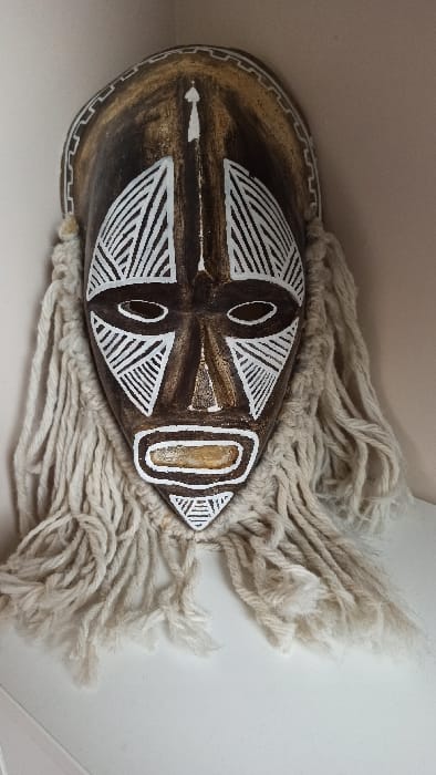 An African mask, primarily brown with white tribal markings, rests sideways. It features circular eye shapes, a rectangular nose section, and a triangular forehead. The mask is surrounded by thick, cream-colored fringe, likely made of yarn or rope, adding a textured contrast to the smooth surface of the mask. The backdrop is a plain, light-colored surface, possibly a wall.