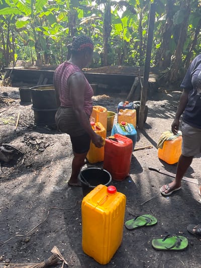 This image depicts an outdoor scene in Aghalokpe, Nigeria, likely related to agricultural processing or commodity handling.

In the foreground, a person, possibly female, is seen from the side, wearing a red and white plaid shirt, dark shorts, and a patterned headwrap. They appear to be actively engaged in an task, standing next to a collection of large, colorful plastic jerrycans and black buckets. Another individual is partially visible in the background, wearing a blue t-shirt and light-colored shorts, also surrounded by similar jerrycans.

The ground is dark and appears to be a mixture of dirt and possibly residue, with scattered organic debris. Several large plastic jerrycans, in shades of yellow, red, blue, and orange, are prominently featured, suggesting the storage or transport of a liquid commodity, potentially palm oil given the regional context. Empty black buckets are also visible. A machete lies on the ground near some feet, and several pairs of flip-flops, including bright green ones, are scattered on the dark ground.

The setting is characterized by lush, dense tropical vegetation, likely banana trees or similar plants, forming a green backdrop. The scene is brightly lit by natural sunlight, casting strong shadows on the ground, indicating it is daytime, possibly midday. Simple wooden structures are present in the background, consistent with a rustic outdoor working area. No visible text can be discerned on any objects or surfaces. The overall impression is one of active labor or processing in a rural, tropical environment.