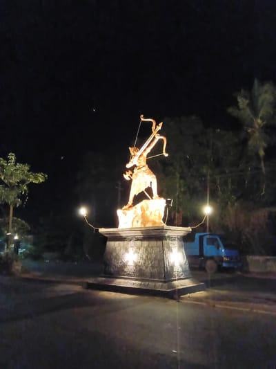 At night in Chopdem, India, a statue of a man with a bow and arrow stands on a raised stone platform. The statue and platform are lit by several lights. Behind the platform is a blue truck. A tree and other greenery are visible in the background. The surrounding area is dark.