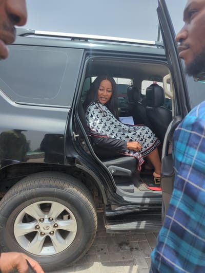 In Unguwan Sanusi, Nigeria, an adult female sits inside the rear passenger seat of a black SUV, the car door is open. She wears a black blazer over a black and white patterned top and has visible dental braces. A man with dark skin and a beard appears in the upper left, close to the car door. Above and to the right, a second man in a blue plaid shirt partially obstructs the view. The ground is paved with gray bricks.