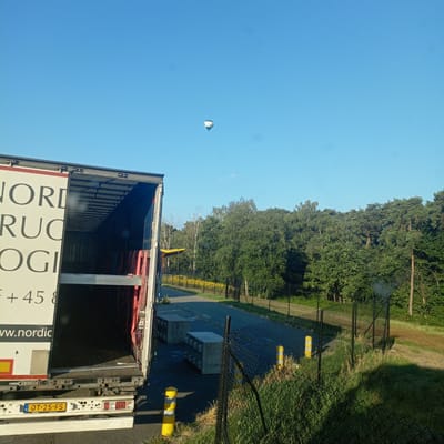 A semi-trailer truck is parked on a paved surface, with its rear doors open, revealing a dark interior. The side of the trailer has text that includes "NORDIC TRUCKING LOGISTICS" and "WWW.NORDIC". A license plate reads "OT-25-FS". Yellow and white bollards are visible along the roadside. In the background, a dense line of trees forms a forest. A hot air balloon is visible in the clear blue sky above the trees. The scene appears to be outdoors, possibly at a logistics facility or rest stop. The location is in Brecht, Belgium.