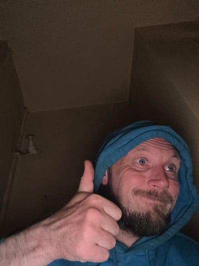 A medium shot shows a Caucasian man with short, light brown hair and a beard, wearing a blue hooded sweatshirt. He is looking up and to his right with wide eyes and a slight smile, his left arm is extended forward, with his thumb up in a "thumbs up" gesture. The man's skin is pale, and his veins are visible on his forearm. The background is a dark, textured wall, with a white pipe fixture visible on the left side of the frame. The image appears to have been taken in a dimly lit, indoor space. There is no indication of the city of Cheltenham, United Kingdom.