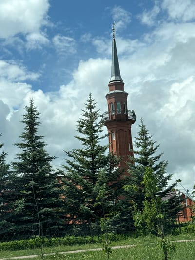 The image displays a red brick minaret of a mosque situated in Belebei, Russia, partially obscured by dense coniferous trees. The minaret features a multi-tiered structure, beginning with a cylindrical red brick base, transitioning to an octagonal section, and culminating in a circular section with a prominent ornate black metal balcony. Within this upper circular section, arched windows with white frames are visible. Above the balcony, a steep, dark grey conical spire ascends, topped with a silver-colored crescent moon finial. Numerous dark green coniferous trees, appearing to be spruce or fir, occupy the midground and foreground, largely concealing the lower sections of the minaret and any adjacent buildings. In the immediate foreground, a strip of green grass with sparse smaller, lighter green deciduous saplings is present. The sky above is predominantly blue, filled with scattered large, white and grey cumulus clouds. No individuals, animals, or vehicles are visible within the frame.