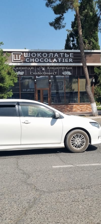 The image shows a white car parked in front of a building with a sign that reads "ШОКОЛАТЬЕ CHOCOLATIER" in large white letters. Below this, in smaller text, it says "Кондитерская Confectionery". The building appears to be a shop, possibly a chocolate shop or confectionery. A tree is growing at an angle in front of the building. The scene is outdoors, with a clear blue sky visible. The car is parked on an asphalt surface, with a white line painted on it, suggesting it's a parking lot. The time of day appears to be daytime, with bright sunlight casting shadows. The overall impression is a casual outdoor scene in a commercial area. The location is identified as Ismoili Somoni, Tajikistan.