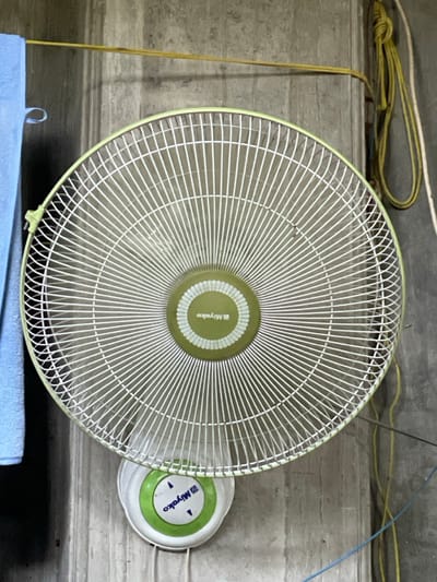 A Miyako brand wall-mounted electric fan is prominently displayed against a concrete wall in Mns Asan, Indonesia. The fan unit consists of a circular light green frame encasing a white protective grille with radial and concentric wires. The central hub of the fan, also light green, features the "Miyako" logo in white. Below the main fan head, the control panel, white with a central green disc and "Miyako" branding, is affixed to the wall, showing two blue arrow indicators. The concrete wall is a uniform grey with vertical striations, indicating a raw or unpainted finish. To the upper left, a light blue towel hangs vertically. To the upper right, a yellow utility rope is secured to the wall with a knot and hangs loosely coiled. A thin yellow string runs horizontally across the top section of the wall. A thin blue electrical wire is visible running vertically down the lower right portion of the wall. No individuals are present.