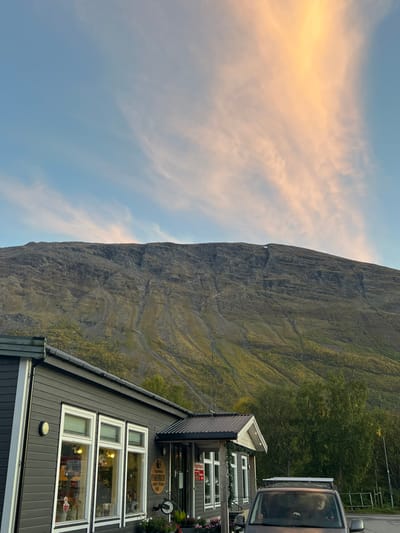 This image depicts a static outdoor scene in Nordkjosbotn - Gárgán, Norway, under a sky with both blue and peach-colored cirrus clouds, indicative of either sunrise or sunset.
In the background, a large, imposing mountain occupies the upper two-thirds of the frame. Its slopes are rugged and textured, displaying exposed grey rock formations, erosion channels, and patches of green and yellowing vegetation, suggesting early autumn or late summer. A small, distinct patch of white material, likely snow or ice, is visible near the mountain's summit, slightly right of the vertical center.
In the mid-ground and foreground, a dark grey, single-story commercial building with horizontal siding is positioned to the left. The building features multiple large rectangular windows with white frames, through which internal lighting is visible, along with reflections. A round brown sign near the building's entrance reads "Stabbursmat AS," with smaller text "Tradisjonell mat fra Nord-Norge." A red rectangular sign is also visible on an adjacent window. The entrance area has a dark corrugated metal awning and is decorated with green garland. Flower boxes containing various plants are situated below the windows. Round light fixtures are mounted on the building's exterior walls.
To the right of the building, a dark-colored van or camper van is partially visible, parked with its front facing towards the viewer. It is equipped with a roof rack that includes what appear to be solar panels. A light-colored animal, resembling a dog, is observable looking out the front passenger window of the vehicle. No human figures are present or clearly identifiable. The immediate ground around the building and vehicle is not fully visible, but some green foliage and darker ground cover are implied.