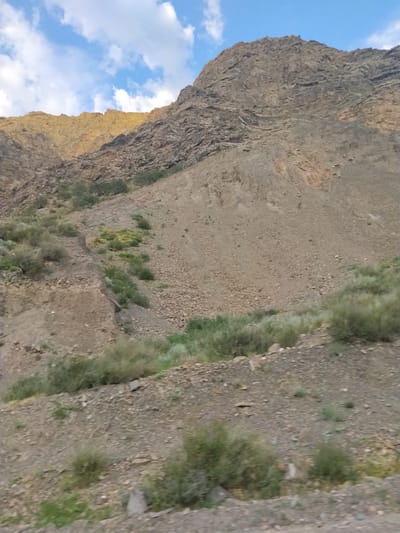 The image depicts a mountainous landscape with sparse vegetation under a partly cloudy sky. The terrain is rocky and arid, with shades of brown and grey dominating the scene. In the foreground, the ground is covered with loose stones and small bushes. The middle ground shows a steep, eroded slope with more scrubby plants and some patches of yellow vegetation. The background features a large, rugged mountain with visible layers of rock. A sliver of yellow light illuminates the upper portion of the mountain, suggesting it is either sunrise or sunset. The image appears to be a landscape photograph taken in a natural environment. There are no people or man-made structures visible in the image, suggesting a remote or undeveloped area.