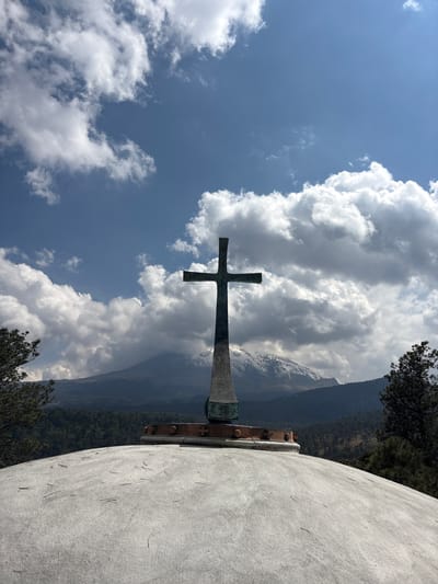 A cross stands in front of clouds and a mountain range. The photograph was taken in San Mateo Ozolco, Mexico. The cross is metallic and mounted on a concrete base. The sky is blue with puffy white clouds. The mountain range is green and brown.