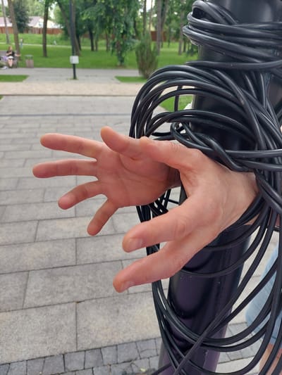 The image shows a close-up of a person's hand, which appears to be trapped or tangled in thick black cables. The cables are coiled around a dark, cylindrical object, and the hand is reaching out, fingers spread, from within the coils. The skin tone of the hand suggests it belongs to a person of light complexion. The background is blurred, but visible elements include a paved walkway made of grey rectangular tiles and green grass with trees, indicating an outdoor setting. In the further background, hints of park benches and distant figures can be seen, suggesting a public park or recreational area. The lighting suggests it is daytime, with natural light illuminating the scene. The overall impression is one of struggle or entrapment due to the cables, though the specific activity or event is not entirely clear. There is no visible text in the image. The context provided indicates the location is Alikonovka, Russia.