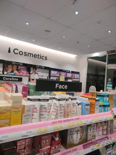 The image shows a retail store interior, likely a pharmacy or beauty store, with shelves stocked with cosmetics and skincare products. A prominent sign reads "Cosmetics" with a lipstick icon. Below this, there are displays featuring different brands like BYS and Maybelline. Further down, "Careline" and "Face" are visible on signs. The shelves are filled with various products, including tubes and bottles with labels such as "Neutrogena Deep Clean," "Collagen," and "LUMINOUS SKIN GLOW." Price tags are visible on some shelves. The lighting appears to be fluorescent from the ceiling tiles. The environment suggests a commercial establishment within Kidapawan City, Philippines, given the context.