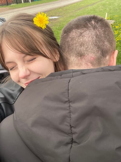 A close-up shot shows two people in Karmaskaly, Russia. A young woman with light brown hair adorned with a yellow dandelion flower is pressing her face against the back of a man's head. The man has short, dark hair. Both are wearing outerwear, with the woman in a black leather jacket and the man in a black puffer jacket. The background is a blurred mix of green grass, yellow flowers, and a paved road.