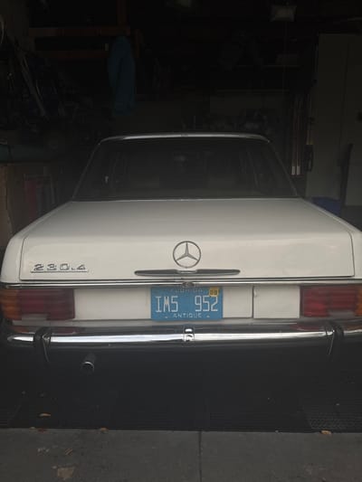 A white Mercedes-Benz 230.4 antique sedan, displaying Florida license plate "IMS 952" with an expiration of "02-28," is parked facing away from the viewer inside a dimly lit garage in Miami Shores, United States of America. The vehicle's rear features a chrome bumper, a centered Mercedes-Benz emblem, and red and amber taillight assemblies. A single exhaust pipe is visible on the lower left. The garage floor consists of dark, possibly rubberized mats over a concrete surface in the foreground. The background reveals a cluttered storage area with various tools, equipment, and a blue garment hanging on a rack or wall, characteristic of a residential garage environment. No individuals are present in the scene.