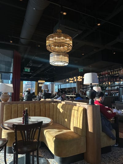 The image portrays the interior of a contemporary restaurant or cafe, situated in Mysiadło, Poland. Natural sunlight enters from large windows on the left, illuminating the space and casting distinct shadows. The ceiling is dark and features exposed industrial ductwork, along with numerous recessed spotlights and two prominent, multi-tiered chandeliers constructed with ribbed, translucent panels.
In the foreground, an L-shaped booth upholstered in mustard-yellow fabric with vertical channel tufting occupies the left side. A round table with a light-colored, possibly marble, top and a dark pedestal base sits before this booth. On the table, a dark glass bottle (likely olive oil), a red and dark salt or pepper grinder, and an empty clear glass are visible. A dark wooden chair with a slatted back is positioned at the table.
Multiple individuals are present throughout the establishment, seated at various tables and booths. On the far right, partially obscured by a table lamp featuring a dark base and light fabric shade, a person with dark hair wearing a red long-sleeved shirt is seated, looking downwards. A dark jacket is draped over their chair. In the background, near the large windows, another individual in a dark winter coat is visible.
To the right of the central seating, a bar or service counter displays the word "CUCINA." Shelves behind this counter are stocked with bottles and glassware. Green "EXIT" signs with directional arrows are affixed to the ceiling. Red curtains frame the windows on the left, which reveal an urban outdoor scene with buildings under a clear sky. The floor throughout the establishment features a patterned mosaic tile design.