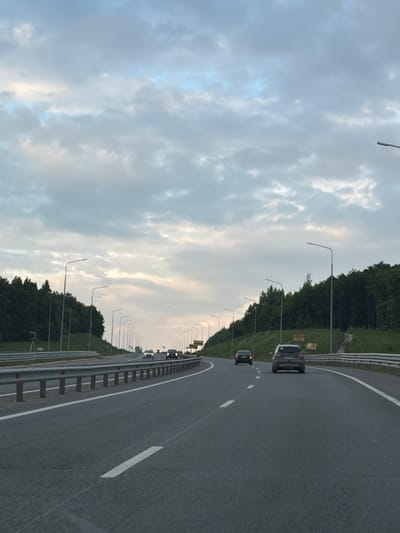 The image shows a view of a highway in Званка, Russia, during the daytime under a cloudy sky. The highway curves to the right, with a line of streetlights and trees lining the left side. Several cars are visible on the road, traveling in the same direction. The road surface is asphalt with white lane markings. In the distance, there are signs on poles, but the text is too small to read. The overall impression is of a typical highway scene on an overcast day.
