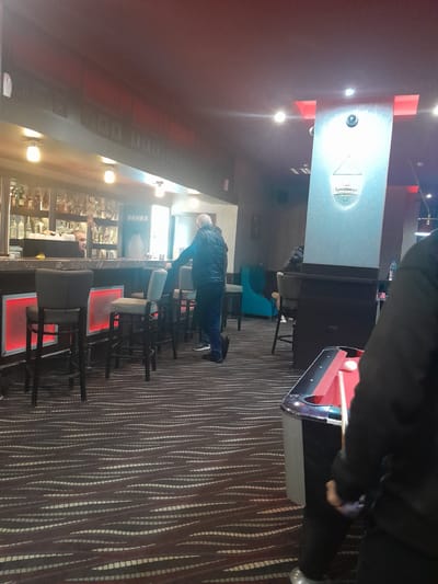 The image depicts the interior of a commercial establishment, identified as "Kamerina" in Topolovgrad, Bulgaria, based on an emblem displayed on a prominent vertical pillar. The space is dimly lit by various artificial light sources, including ceiling-mounted fixtures, sconces above a bar, and red LED panels integrated into the bar's facade. The floor is covered with a dark carpet featuring a distinctive wavy pattern of light-colored, segmented lines.
Four individuals are present. In the immediate foreground on the right, a person dressed in dark clothing holds a pool cue, positioned near a red-felted pool table, indicating an intention to play. A white cue ball is visible on the table. To the left, in the mid-ground, a male with short or receding grey hair, wearing a dark puffer vest over a long-sleeved shirt and dark pants, stands leaning against a dark bar counter. He is oriented towards a male bartender, whose head and upper torso are visible behind the counter amidst shelves stocked with numerous bottles of alcohol. A white sign with the Cyrillic word "БАНКЯ" is present behind the bar. The bar counter is fronted by multiple dark, high-backed stools, some featuring red-lit panels. A tall, light grey vertical pillar with red accents near the top and a circular object (possibly a camera or speaker) is situated to the right of the bar. In the background, beyond the pillar, a fourth individual is partially visible, seated in a dark chair, near a turquoise armchair. The primary actions observed are one person preparing to play pool and another standing at the bar, presumably awaiting service from the bartender.