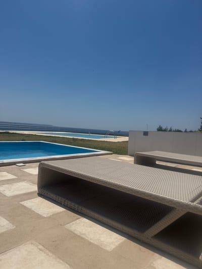 The image depicts an outdoor scene with two rectangular swimming pools and lounge chairs. The foreground is dominated by a beige wicker lounge chair. Beyond this, two swimming pools with clear blue water are visible. In the background, a low white fence separates the area from a body of water, likely the sea, with a faint horizon line. The sky is a clear, cloudless blue. The location is identified as Nea Moudania, Greece.
