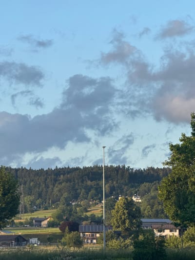 This image depicts a rural landscape, set in Fehraltorf, Switzerland, under a partly cloudy sky. A dominant feature is a densely forested hillside or mountain range stretching horizontally across the mid-ground, covered in vibrant green trees. Above this, the sky is a soft blue with scattered grey and white clouds, indicating a mix of clear patches and cloud cover.
Several buildings are visible, situated both within the tree line on the hillside and in the lower foreground. These include houses, some with reddish roofs and white walls, while others, particularly in the lower right, feature solar panels on their roofs, suggesting modern dwellings or agricultural structures.
A prominent, tall, slender white pole stands vertically in the left-center, reaching towards the sky. A small, dark object, likely a bird, is perched at its very top. In the lower right foreground, a yellow and black diamond-shaped sign is partially visible, indicating a road or pathway in the vicinity. Green fields or cultivated land can also be seen in the lower right, adjacent to the buildings and trees.
The scene is captured during the daytime, possibly in the late afternoon, with soft, ambient lighting. There is no explicit human activity or visible text in the image. The overall impression is one of a peaceful, verdant landscape with subtle signs of human presence.